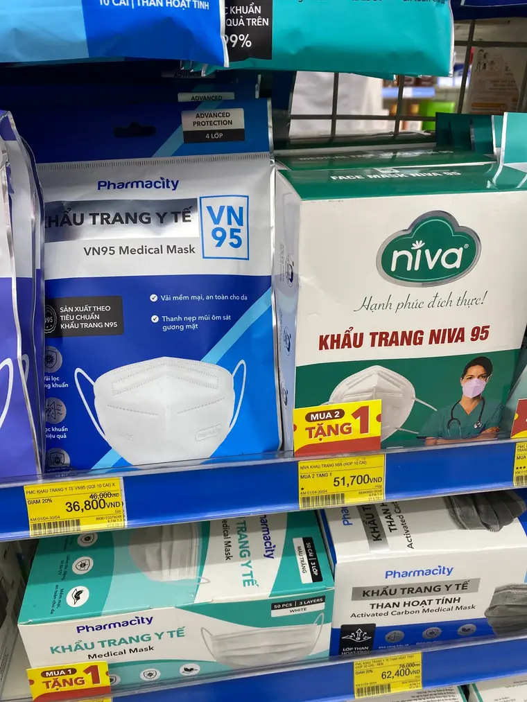 Полка аптеки Pharmacity. Слева синий пакет «KHẨU TRANG Y TẾ VN95 / VN95 Medical Mask» с белой маской на картинке, справа зелёная коробка «KHẨU TRANG NIVA 95» с врачом в маске и стетоскопом. Жёлтые ценники на бортике полки: 36 800 донгов вместо зачёркнутых 46 000 («GIẢM 20%») и 51 700 донгов. На коробке Niva жёлтая наклейка «MUA 2 TẶNG 1», а на нижней полке у коробки Pharmacity — «MUA 1 TẶNG 1» и ценник 62 400 вместо 78 000.