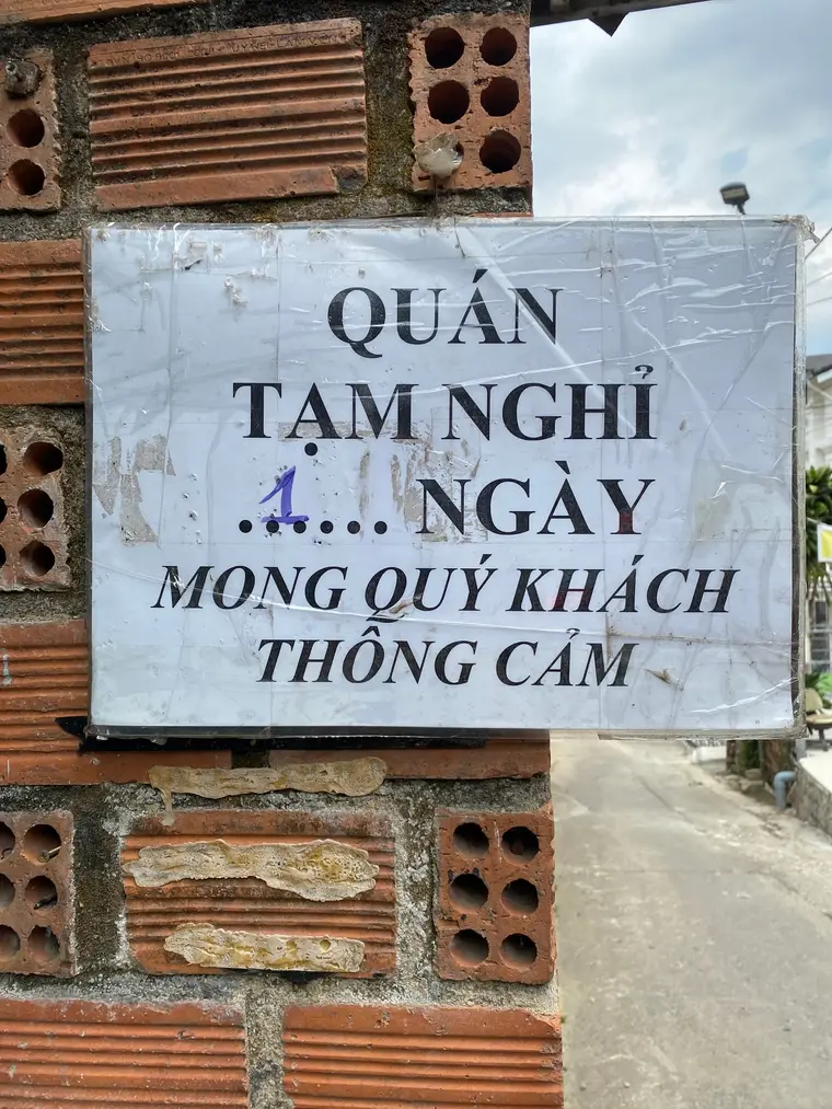 Заламинированная табличка на столбе из красного кирпича: «QUÁN TẠM NGHỈ …1… NGÀY MONG QUÝ KHÁCH THÔNG CẢM». Единица вписана в пропуск фиолетовой ручкой. Справа уходит пустая улица.