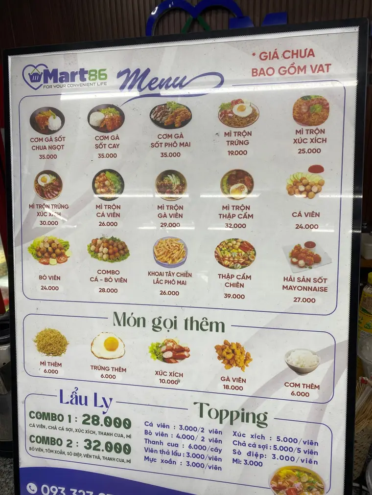 Печатное меню в рамке на подставке. Слева логотип «Mart86», по центру «Menu». Три ряда фотографий блюд, под каждой название по-вьетнамски и цена в донгах — Cơm gà sốt chua ngọt 35.000, Mì trộn trứng 19.000, Bò viên 24.000. Ниже раздел «Món gọi thêm» с добавками по 6.000–18.000, под ним комбо «Lẩu Ly» и столбики цен на топпинги. В правом верхнем углу красным: «* GIÁ CHƯA BAO GỒM VAT».