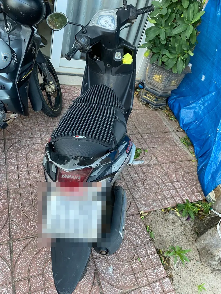 Чёрный скутер Yamaha стоит на красной тротуарной плитке у двери дома, снят сверху сзади: сиденье в массажной накидке из чёрных резиновых шариков, задний пластик потрескался и облез до белого, номерной знак замазан пикселями; слева впритык стоит второй байк, справа синий брезент и кадка с растением.