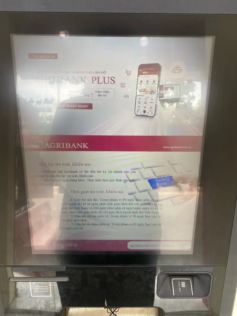 Экран банкомата Agribank за стеклом, в котором отражается улица. Верх экрана занимает реклама мобильного приложения: «AGRIBANK E-MOBILE BANKING — PHIÊN BẢN MỚI», крупно «AGRIBANK PLUS», кнопка «CẬP NHẬT NGAY» и фотография телефона с открытым приложением. Ниже красная полоса с логотипом банка и адресом сайта, под ней вьетнамский текст с заголовками «Thủ tục tra soát, khiếu nại» и «Thời gian tra soát, khiếu nại» — порядок и сроки разбирательства по спорной операции, с пунктами про 80 и 160 дней. В самом низу «GIAO DỊCH KHÔNG DÙNG THẺ (CARDLESS)».