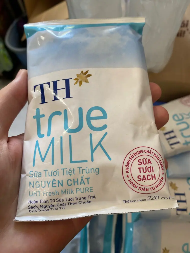 Рука держит мягкую упаковку молока TH true MILK на 220 мл. Надпись на ней: «Sữa Tươi Tiệt Trùng NGUYÊN CHẤT», ниже «UHT Fresh Milk PURE», справа круглая красная печать «Sữa Tươi Sạch». Позади лежат такие же пачки и прозрачный пакет.