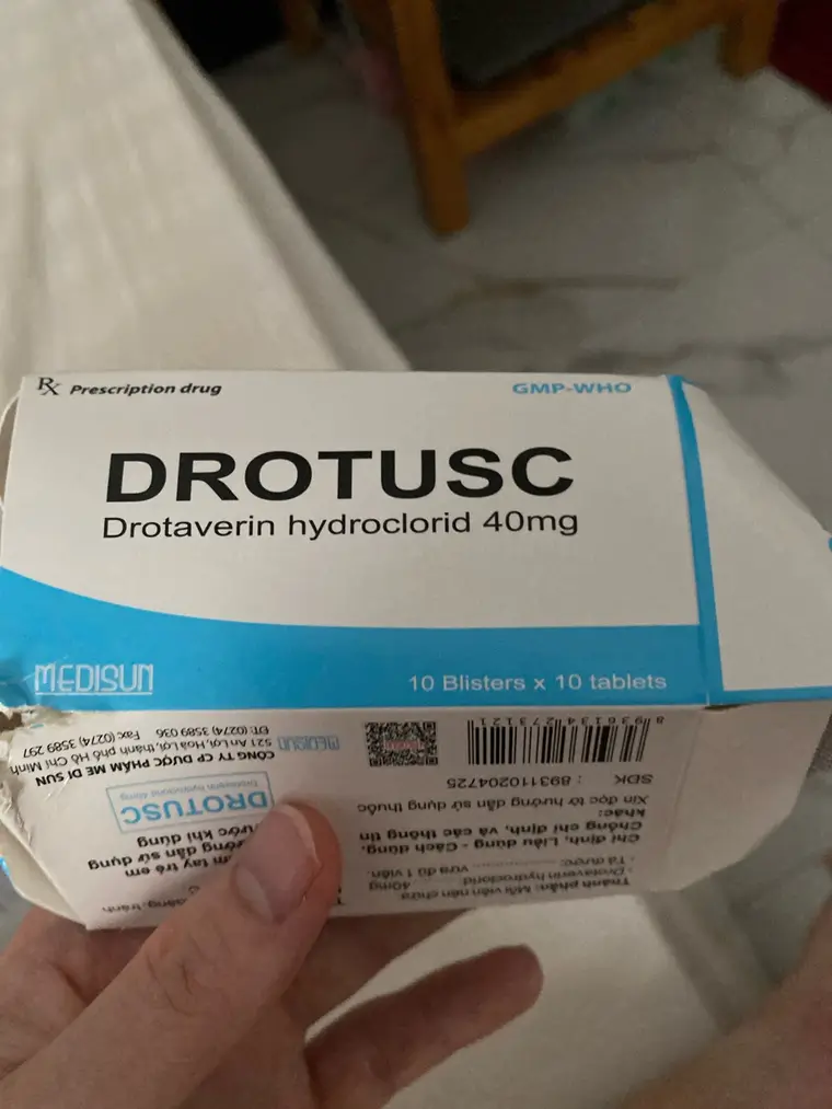 Рука держит картонную коробку лекарства. Сверху мелко «Rx Prescription drug» и «GMP-WHO», под ними крупно «DROTUSC», строкой ниже — «Drotaverin hydroclorid 40mg». На синей полосе марка «MEDISUN» и «10 Blisters x 10 tablets». Боковая грань повёрнута к зрителю вверх ногами: вьетнамская инструкция мелким шрифтом, штрихкод и номер регистрации. Позади белое покрывало и деревянный стул.
